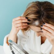 Woman in white robe checking her hair for gray strands using a mirror on blue background.