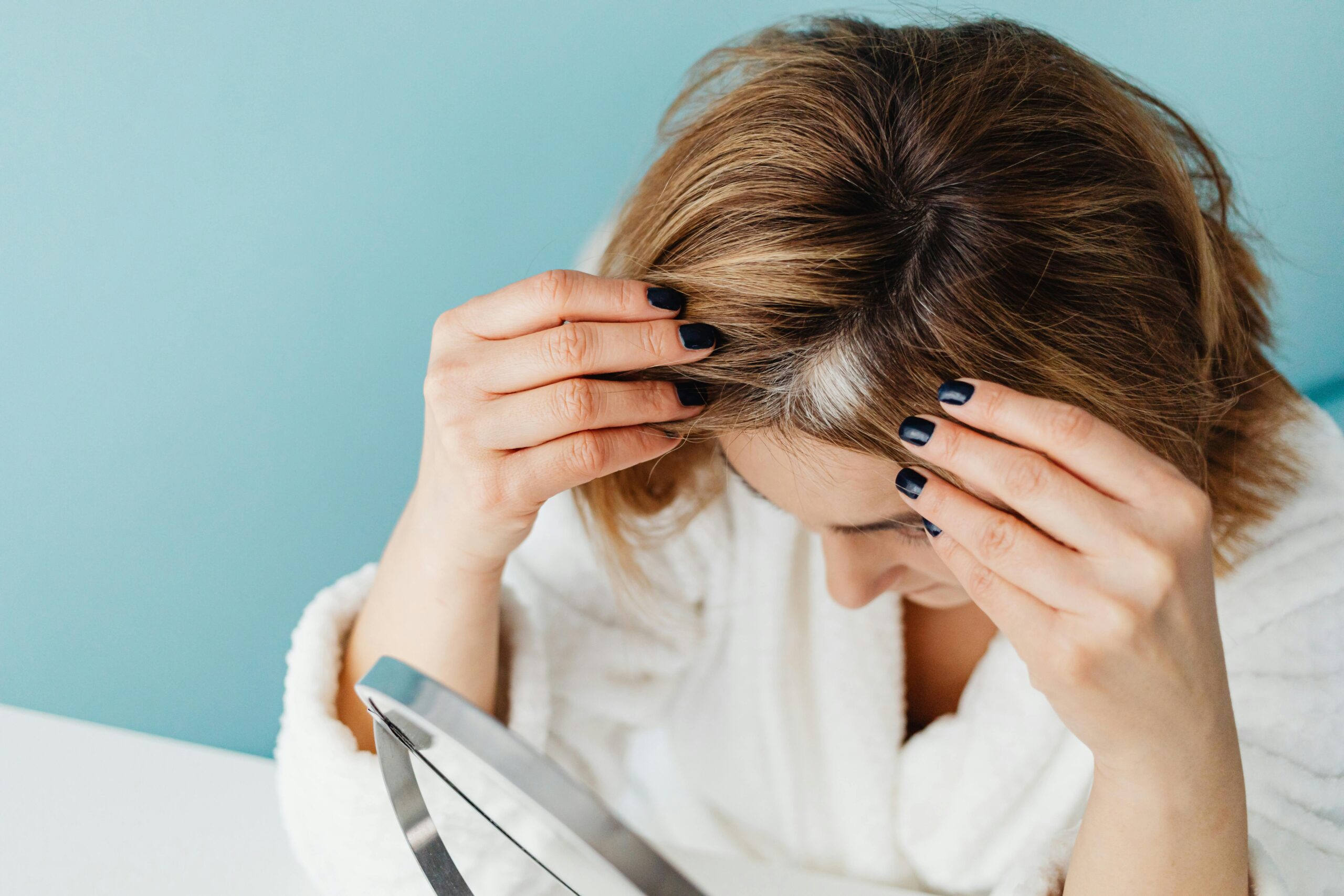 Woman in white robe checking her hair for gray strands using a mirror on blue background.